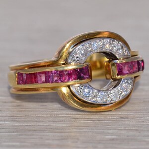 May include: A gold ring with a distinctive design. The ring features a central circle of small, clear stones, with a band of rectangular, red stones on either side. The ring has a two-tone gold and silver appearance.