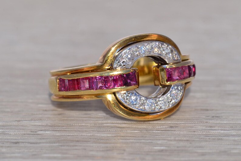 May include: A gold ring with a distinctive design. The ring features a central circle of small, clear stones, with a band of rectangular, red stones on either side. The ring has a two-tone gold and silver appearance.