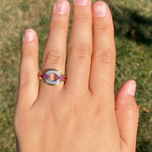 May include: A gold ring featuring an oval centrepiece with small white stones, framed by red stones. The ring is displayed on a hand, set against a blurred green backdrop.