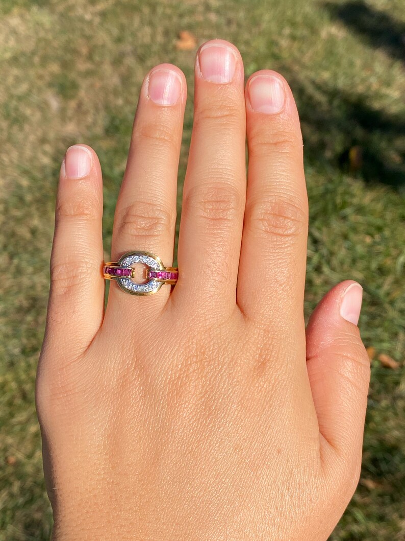 May include: A gold ring featuring an oval centrepiece with small white stones, framed by red stones. The ring is displayed on a hand, set against a blurred green backdrop.