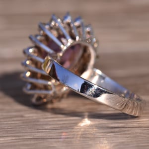 May include: A silver ring featuring a large purple gemstone encircled by smaller clear gemstones. The ring is set in a halo style.