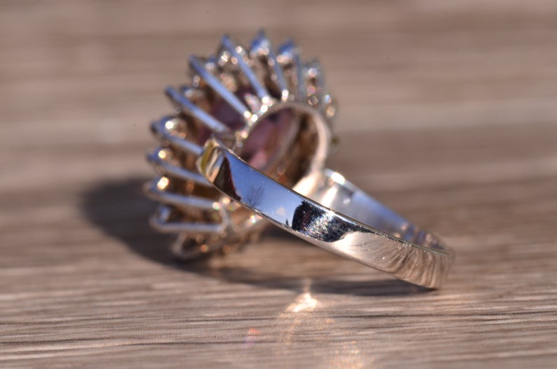 May include: A silver ring featuring a large purple gemstone encircled by smaller clear gemstones. The ring is set in a halo style.