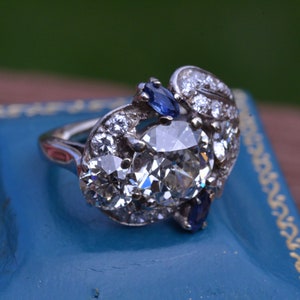 May include: A silver ring featuring a large, oval-shaped diamond surrounded by smaller diamonds and two blue sapphires.