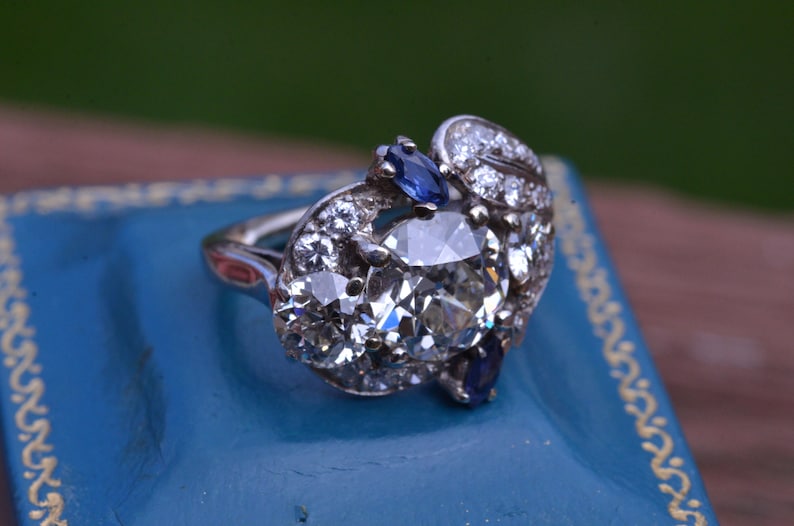 May include: A silver ring featuring a large, oval-shaped diamond surrounded by smaller diamonds and two blue sapphires.