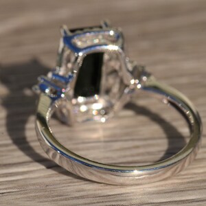 K&ouml;nnte beinhalten: Ein silberner Ring mit einem gro&szlig;en schwarzen Edelstein, der von kleineren klaren Edelsteinen umgeben ist. Der Ring ist in einer Krappenfassung gefasst und hat eine polierte Oberfl&auml;che.