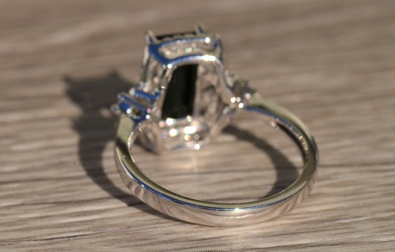 K&ouml;nnte beinhalten: Ein silberner Ring mit einem gro&szlig;en schwarzen Edelstein, der von kleineren klaren Edelsteinen umgeben ist. Der Ring ist in einer Krappenfassung gefasst und hat eine polierte Oberfl&auml;che.