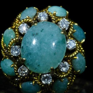 Könnte beinhalten: Ein goldener Ring mit einem großen ovalen grünen Edelstein, umgeben von kleineren grünen Edelsteinen und Diamanten.