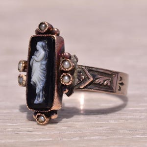 Pu&ograve; includere: Anello antico con cammeo nero rettangolare raffigurante una figura classica bianca. L'anello ha una fascia dorata con dettagli decorativi e piccoli accenti di perle. Un gioiello vintage.