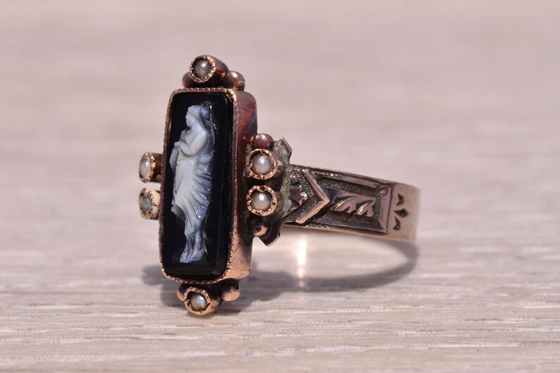 Pu&ograve; includere: Anello antico con cammeo nero rettangolare raffigurante una figura classica bianca. L'anello ha una fascia dorata con dettagli decorativi e piccoli accenti di perle. Un gioiello vintage.