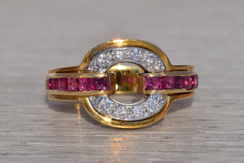 May include: A gold ring with a distinctive design, featuring a central open circle. The ring is embellished with sparkling white diamonds and vibrant red gemstones. The gemstones are set in a channel setting, enhancing the ring's elegance.