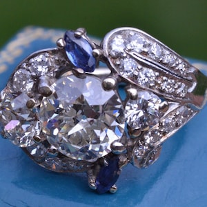 May include: A silver ring featuring a large, round, clear diamond surrounded by smaller diamonds. The ring also has two blue sapphires on either side of the main diamond.
