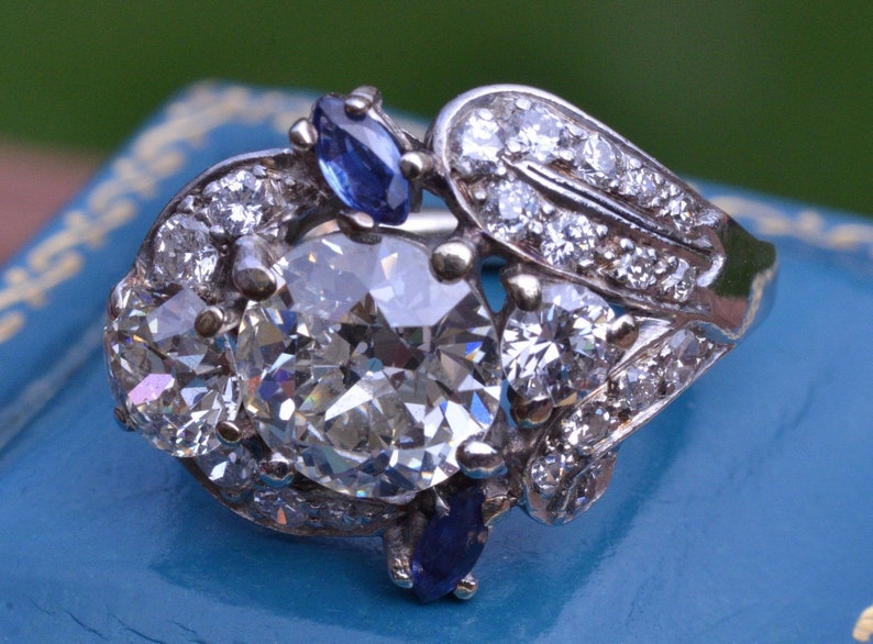 May include: A silver ring featuring a large, round, clear diamond surrounded by smaller diamonds. The ring also has two blue sapphires on either side of the main diamond.