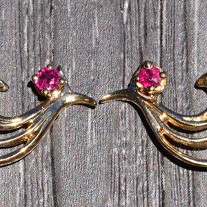 May include: A pair of gold earrings with a stylized bird design and a pink gemstone in the center of each earring.