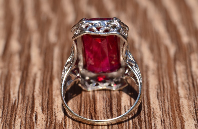 May include: A silver ring featuring an emerald-cut red gemstone set in a filigree design.