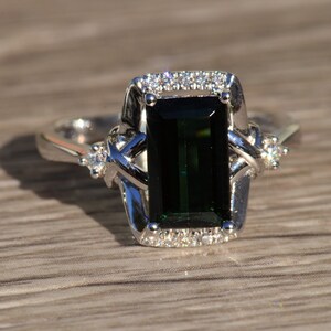 K&ouml;nnte beinhalten: Ein silberner Ring mit einem gro&szlig;en smaragdgr&uuml;nen Edelstein, der von kleineren Diamanten umgeben ist. Der Ring hat ein zartes, filigranes Design.