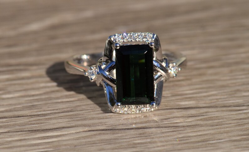 K&ouml;nnte beinhalten: Ein silberner Ring mit einem gro&szlig;en smaragdgr&uuml;nen Edelstein, der von kleineren Diamanten umgeben ist. Der Ring hat ein zartes, filigranes Design.