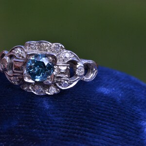 K&ouml;nnte beinhalten: Ein silberner Ring mit einem blauen Edelstein, der von kleineren Diamanten umgeben ist. Der Ring liegt auf einem blauen Samtkissen.