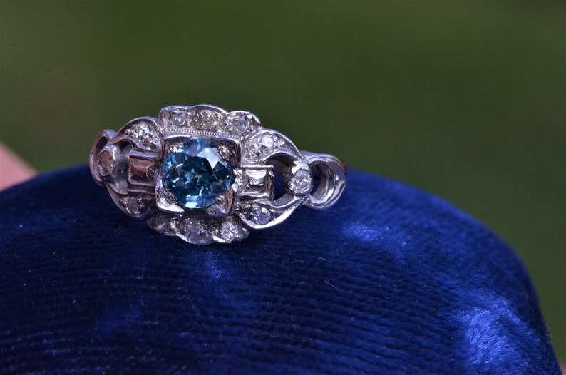 K&ouml;nnte beinhalten: Ein silberner Ring mit einem blauen Edelstein, der von kleineren Diamanten umgeben ist. Der Ring liegt auf einem blauen Samtkissen.