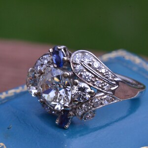 May include: A vintage engagement ring featuring a large, round, clear diamond surrounded by smaller diamonds and two blue sapphires. The ring is set in white gold and has a delicate, floral design.