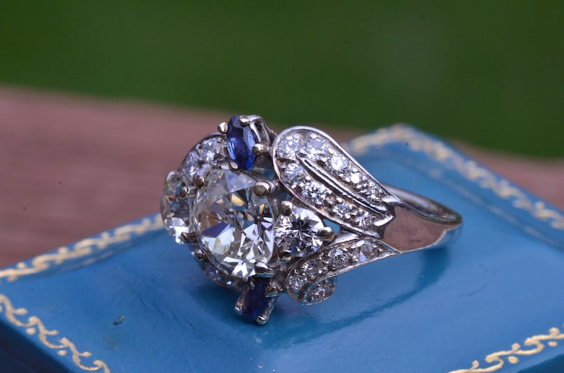 May include: A vintage engagement ring featuring a large, round, clear diamond surrounded by smaller diamonds and two blue sapphires. The ring is set in white gold and has a delicate, floral design.