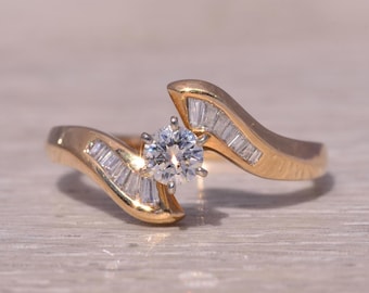 May include: A gold ring with a single round diamond and smaller baguette diamonds set in the band. The ring has a unique, curved design.