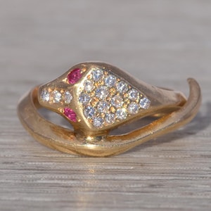 Op de afbeelding: Een goudkleurige slangenring met een kop bezet met kleine, heldere stenen en twee rode stenen als ogen. De ring heeft een opgerold ontwerp, waarbij het lichaam van de slang de band vormt. De ring ligt op een lichtgekleurd, getextureerd oppervlak.