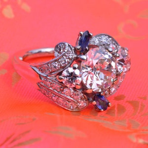 May include: A silver ring featuring a large, round, clear diamond surrounded by smaller diamonds and two blue sapphires.