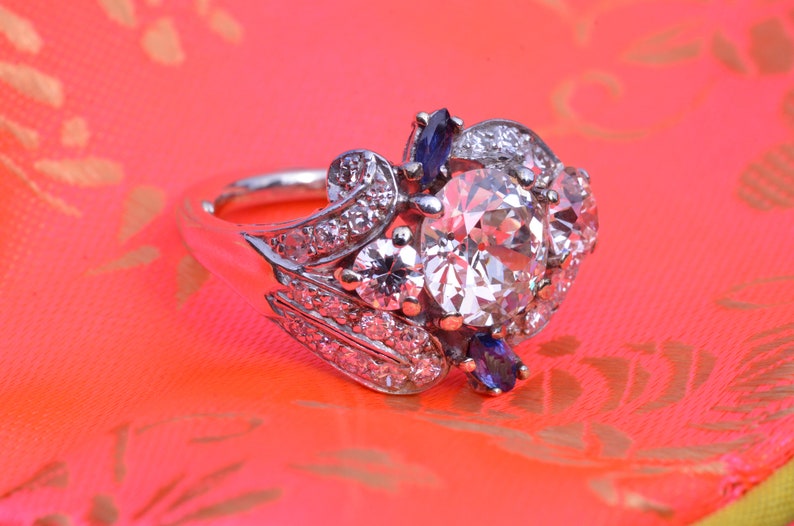 May include: A silver ring featuring a large, round, clear diamond surrounded by smaller diamonds and two blue sapphires.