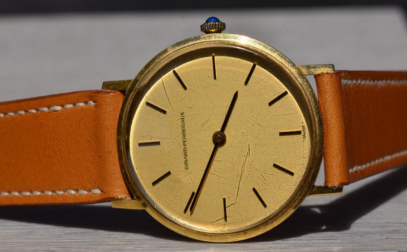 May include: A gold wristwatch with a brown leather band. The watch face is gold with black Roman numeral hour markers and a scratch-resistant crystal. The watch face is signed "Girard-Perregaux".