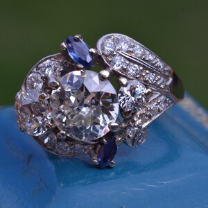 May include: A silver ring featuring a large, round, clear diamond surrounded by smaller diamonds. The ring also has two blue sapphires on either side of the centre diamond.