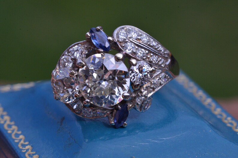 May include: A silver ring featuring a large, round, clear diamond surrounded by smaller diamonds. The ring also has two blue sapphires on either side of the centre diamond.
