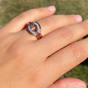 May include: A gold ring with a distinctive design. The ring has a circular centrepiece with small white stones and a band of red stones. The ring is set against a blurred green background.