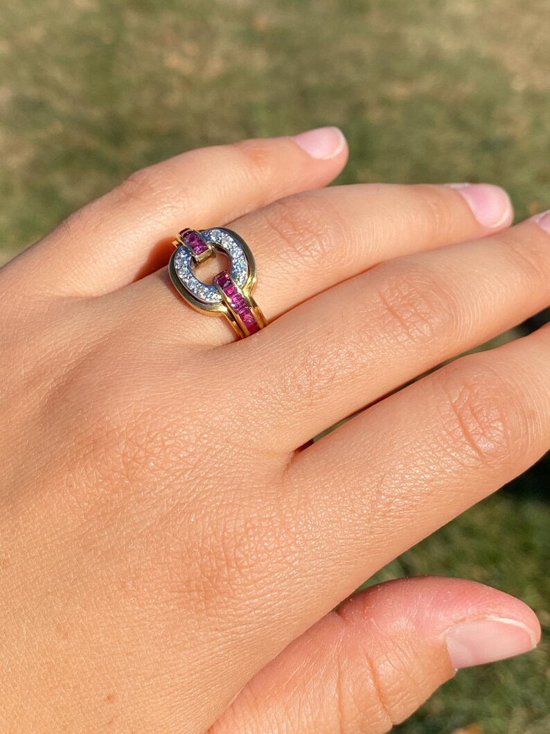 May include: A gold ring with a distinctive design. The ring has a circular centrepiece with small white stones and a band of red stones. The ring is set against a blurred green background.
