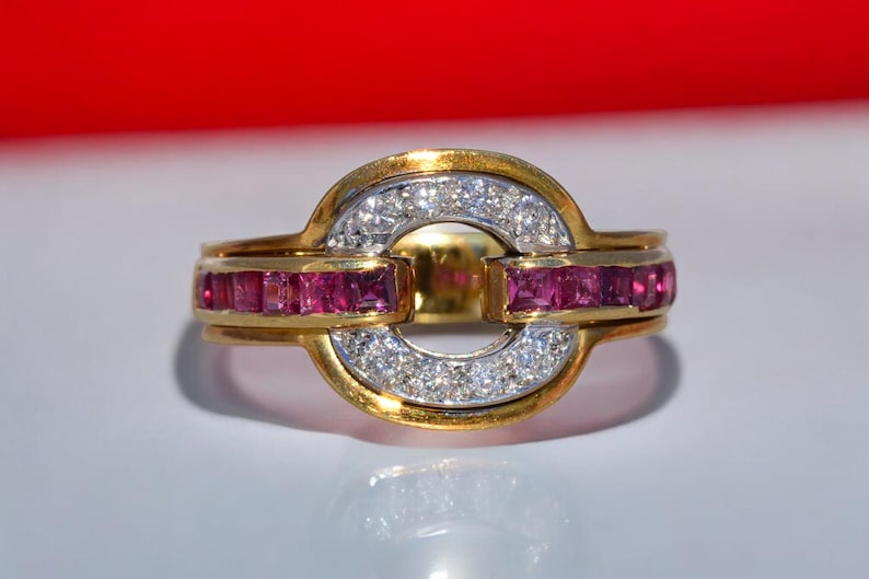 May include: A gold ring with a central oval design. The ring features a band of small, sparkling diamonds and a row of square-cut rubies. The ring has a polished gold finish and a bright, eye-catching appearance.