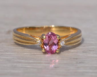 Può includere: Un anello d'oro con una gemma rosa ovale come elemento centrale. L'anello ha una fascia a coste e piccoli diamanti ai lati della pietra principale. L'anello è posto su una superficie di legno chiaro.