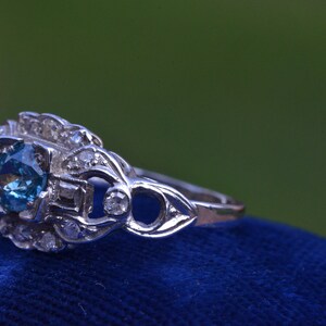 K&ouml;nnte beinhalten: Ein silberner Ring mit einem blauen Edelstein, der von kleineren Diamanten umgeben ist. Der Ring liegt auf einem blauen Samt-Hintergrund.