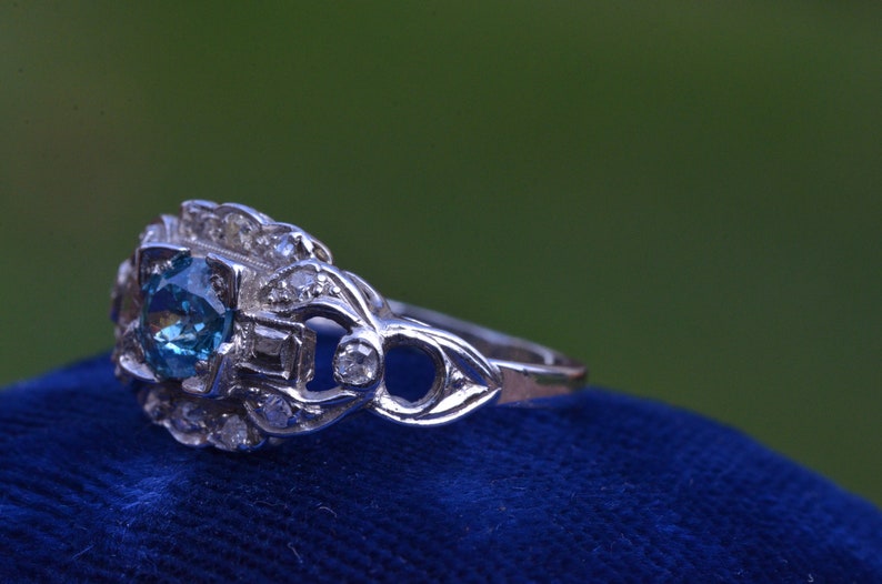 K&ouml;nnte beinhalten: Ein silberner Ring mit einem blauen Edelstein, der von kleineren Diamanten umgeben ist. Der Ring liegt auf einem blauen Samt-Hintergrund.