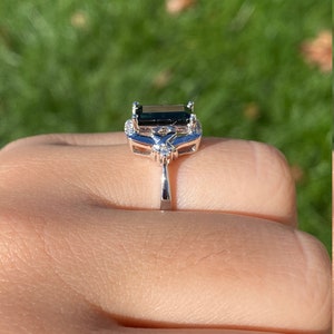 K&ouml;nnte beinhalten: Ein silberner Ring mit einem dunkelgr&uuml;nen Smaragd-Schliff-Edelstein, umgeben von kleineren klaren Edelsteinen.