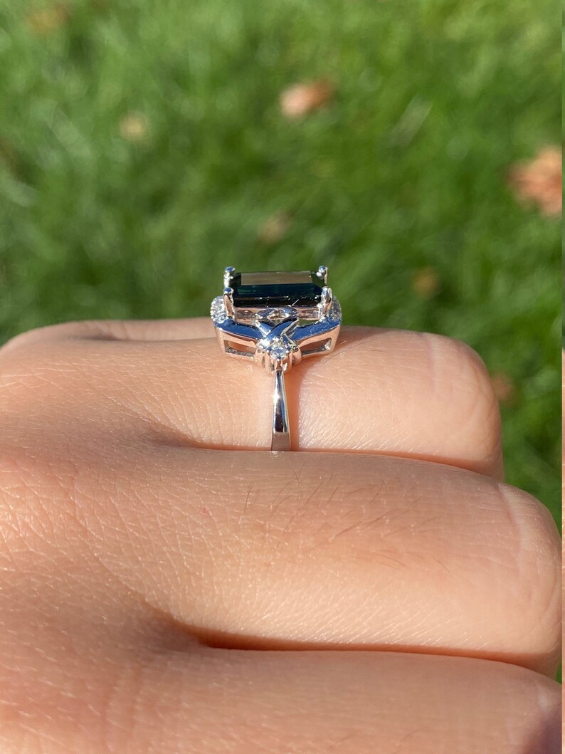 K&ouml;nnte beinhalten: Ein silberner Ring mit einem dunkelgr&uuml;nen Smaragd-Schliff-Edelstein, umgeben von kleineren klaren Edelsteinen.