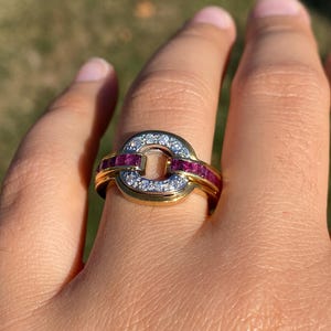 May include: A gold ring with a circular design. The ring features a row of small, sparkling white stones and a row of red stones. The ring is on a finger.