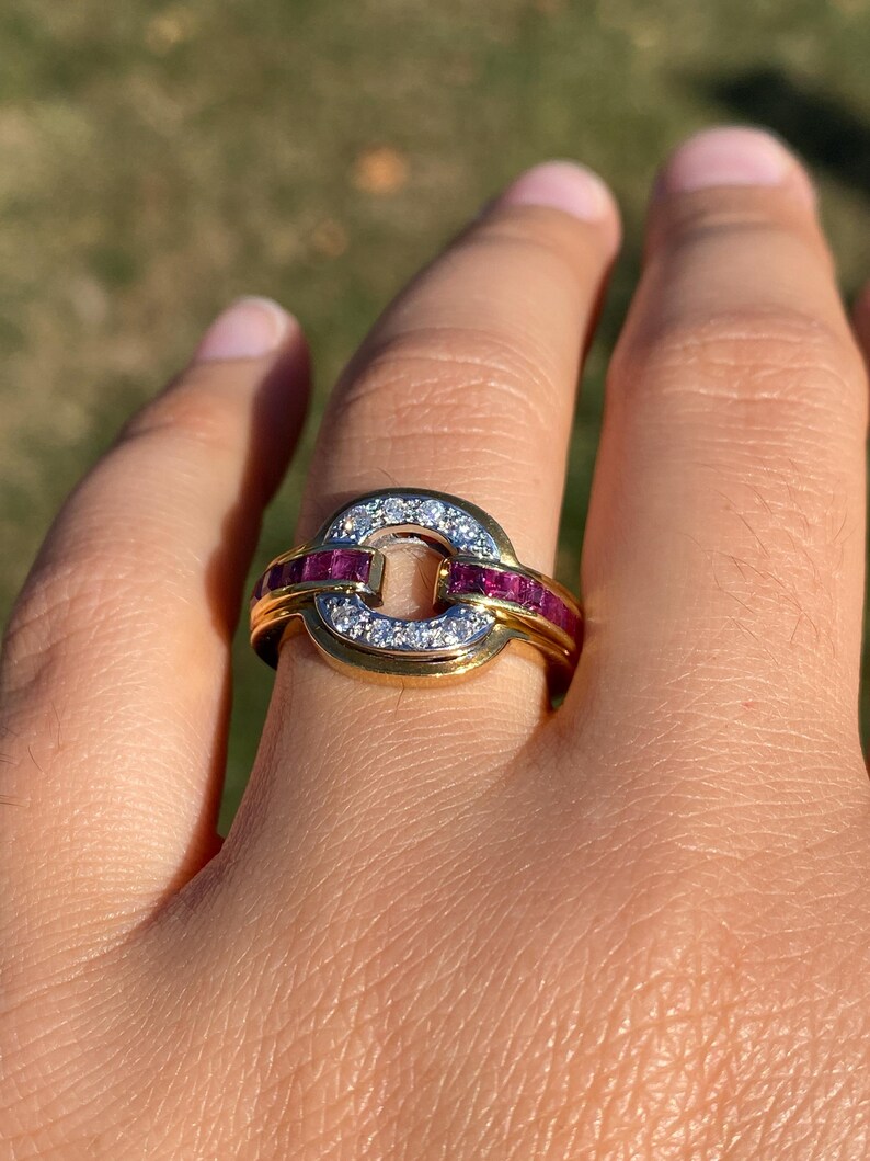 May include: A gold ring with a circular design. The ring features a row of small, sparkling white stones and a row of red stones. The ring is on a finger.