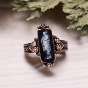 Pu&ograve; includere: Anello in stile antico con un elemento centrale rettangolare in onice nero con un cammeo bianco di una figura classica. L'anello ha una fascia color bronzo con accenti decorativi e piccole perle.