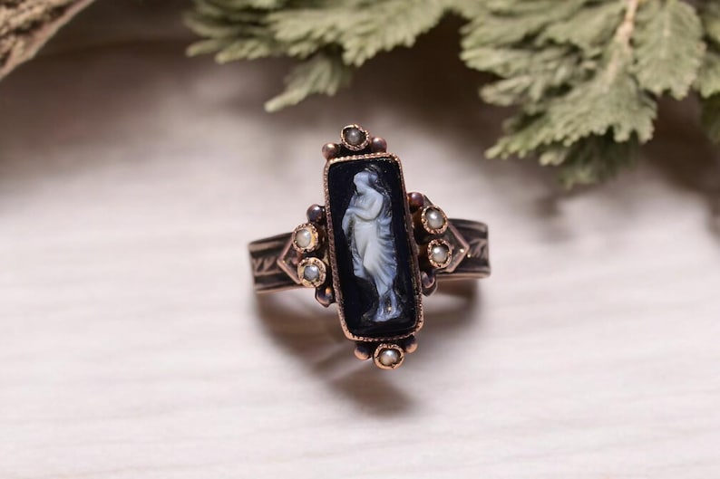 Pu&ograve; includere: Anello in stile antico con un elemento centrale rettangolare in onice nero con un cammeo bianco di una figura classica. L'anello ha una fascia color bronzo con accenti decorativi e piccole perle.