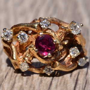 Può includere: Un anello d'oro con una pietra preziosa di rubino circondata da diamanti più piccoli. L'anello ha un design unico e organico che ricorda i rami.