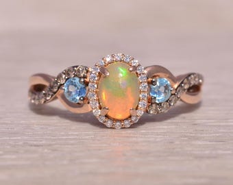 May include: Rose gold ring featuring an oval opal center stone surrounded by a halo of small diamonds. Two blue gemstones flank the opal, set within a twisted band with additional diamond accents. A beautiful piece of jewelry.