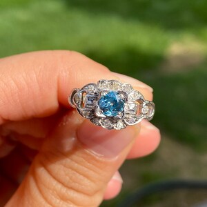 K&ouml;nnte beinhalten: Ein silberner Ring mit einem blauen Edelstein, der von kleineren Diamanten umgeben ist. Der Ring hat ein zartes, filigranes Design.