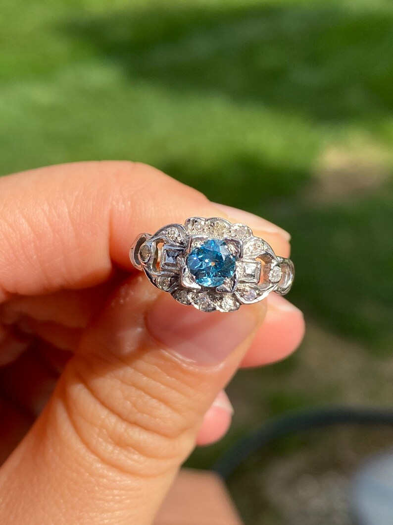K&ouml;nnte beinhalten: Ein silberner Ring mit einem blauen Edelstein, der von kleineren Diamanten umgeben ist. Der Ring hat ein zartes, filigranes Design.