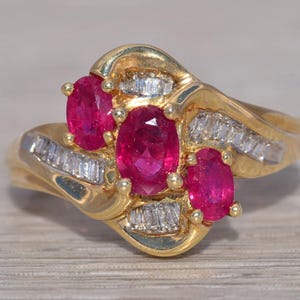 Könnte beinhalten: Ein goldener Ring mit drei ovalen Rubin-Edelsteinen und einer Reihe von Baguette-geschliffenen Diamanten.