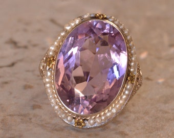May include: A ring with a large oval amethyst gemstone surrounded by a border of small pearls and gold accents.