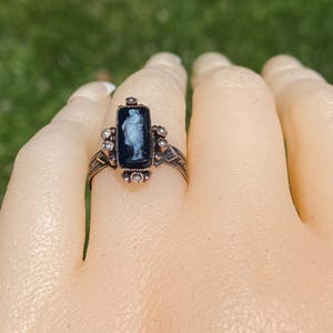 Pu&ograve; includere: Un anello vintage con un cammeo nero rettangolare con una figura bianca. L'anello ha una fascia in metallo color bronzo con accenti decorativi e piccole pietre trasparenti. L'anello &egrave; posto su una mano.